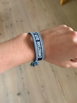 Blått Christian Dior tygarmband - Snyggt blått tygarmband från Christian Dior med vit brodyr och text. Perfekt accessoar för att lyfta din outfit med en exklusiv touch.
