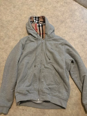 Burberry zip hoodie - Snygg grå hoodie med dragkedja och fickor framtill. Huvan har ett klassiskt rutigt mönster på insidan och justerbara snören. Perfekt för en chill och avslappnad stil. Har lite rosa färg på sig på framsidan men tror att det ska kunna gå bort