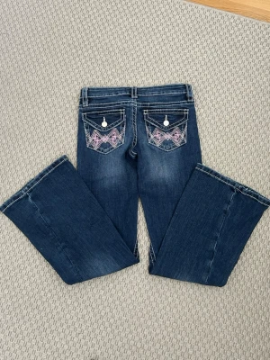 Mörkblå bootcut jeans med brodyr - Snygga mörkblå bootcut jeans med kontrastsömmar och broderade detaljer i rosa och vitt på bakfickorna. Jeansen har låg midja och klassisk femficksmodell. Perfekta för dig som gillar en avslappnad men trendig stil.