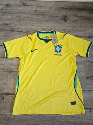 Gul Brasilien fotbollströja 2026 - Snygg gul Brasilien fotbollströja från Nike med blå och gröna detaljer. Tröjan har korta ärmar, rund halsringning och Brasilien-emblemet på bröstet. Perfekt för dig som vill stötta landslaget eller använda till träning.