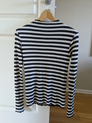 Randig ribbad långärmad topp - Säljer en svartvit randig topp med ribbad struktur och långa ärmar. Toppen har en smal passform och lätt vågiga kanter vid ärmslut och nederkant. Perfekt att styla med jeans eller kjol för en clean look.