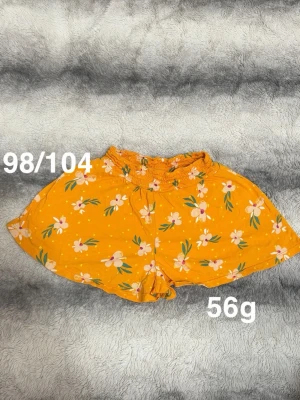 Oranga  blommiga shorts med resårmidja - Säljer ett par oranga shorts med vita och rosa blommor samt gröna blad. Shortsen har en bekväm resår i midjan och är tillverkade i ett lätt och luftigt tyg, perfekta för varma dagar. Passar dig som gillar färgglada och somriga plagg.🌸 Fint använt skick. Storlek 98/104 