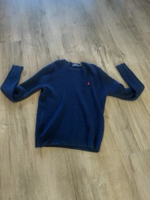 Mörkblå kabelstickad tröja - Mörkblå kabelstickad tröja med rund hals och långa ärmar. Tröjan har ett diskret rött broderat märke på bröstet och ribbade muddar vid ärmslut och nederkant. Perfekt för dig som gillar klassisk och stilren look.