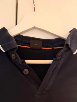 Svart pikétröja från BOSS - Svart pikétröja från BOSS med korta ärmar och vit kant på krage och ärmslut. Klassisk passform med diskret logotyp på bröstet. Perfekt till jeans eller shorts för en clean look.