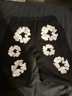 Svarta shorts med vita blommor - Svarta shorts med elastisk midja och dragsko. Dekorerade med stora vita blommönster på både fram- och baksidan. Perfekta för en avslappnad och trendig stil. Materialet ser ut att vara bomull eller bomullsblandning.