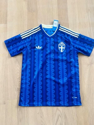 Blå Sverige borta vm tröja - Säljer en blå fotbollströja från Adidas med Sveriges landslagsemblem på bröstet. Tröjan har ett retroinspirerat mönster, vita Adidas-ränder på axlarna och V-ringad krage med vit kant. Tillverkad i lätt och ventilerande material, perfekt för träning eller match.