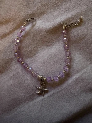 Lila pärlarmband med sjöstjärna, köpt i Thailand - Sött armband med facetterade lila pärlor och små silvriga detaljer. Armbandet har en silverfärgad sjöstjärneberlock som hänger i mitten och justerbart spänne. Perfekt accessoar för dig som gillar strandvibbar och pastellfärger.