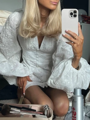 Adoore klänning  - Säljer min superfina adoore klänning i modellen Florence dress. Supersnygg vit kortklänning med stora puffärmar och knappar framtill. Klänningen har v-ringning och ett subtilt blommigt mönster i tyget. Är i superfint skick och endast använd ett fåtal gånger. 
