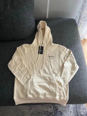 Krämvit hoodie Hampton 44 - Snygg krämvit hoodie från Hampton 44 med tryck på bröstet och ryggen. Hoodien har stor huva, dragsko och en klassisk magficka. Tillverkad i mjukt material som känns skönt mot huden. Perfekt för en avslappnad och trendig look.
