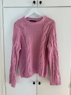 Rosa kabelstickad tröja - Säljer en rosa kabelstickad tröja med rund halsringning och breda ribbade muddar vid ärmslut och nederkant. Tröjan har ett klassiskt flätmönster längs ärmar och framstycke, vilket ger en trendig och mysig look.