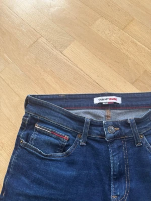 Tommy Jeans  - Snygga mörkblå jeans från Tommy Jeans med klassisk femficksmodell och raka ben. Jeansen har subtila slitningar och röda detaljer vid myntfickan. Jeansen är i storlek 31W 32L och är mer raka i benen än slim. Jeansen är i mycket bra skick då de typ aldrig använts.
