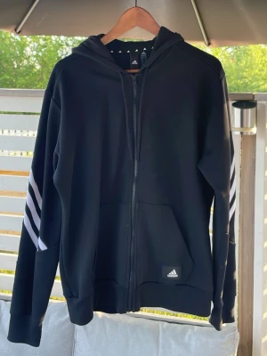 Svart hoodie med dragkedja från Adidas - Säljer en svart hoodie från Adidas med dragkedja framtill och klassiska vita ränder längs ärmarna. Tröjan har huva med snörning och två fickor fram. Adidas-logga på vänster sida nedtill. Perfekt för en sportig och avslappnad stil.