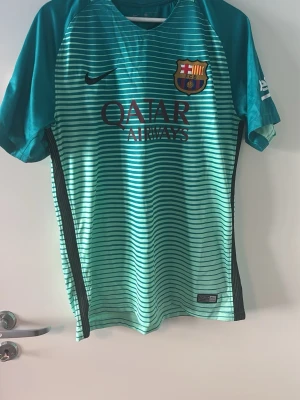 Barcelona tröja  - RETRO BARCA TRÖJA. Säljer en turkos FC Barcelona matchtröja från Nike med Dri-FIT material. Tröjan har tunna horisontella ränder, klubbmärke och Qatar Airways-logga på bröstet samt svarta detaljer längs sidorna. Perfekt för fotbollsträning eller som supporterplagg.