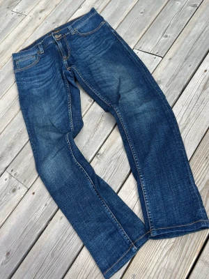 Blå jeans från Nudie Jeans Co W30 L30 - Säljer ett par klassiska blå jeans från Nudie Jeans Co i storlek W30 L30. Jeansen har raka ben, fem fickor och snygga slitningar framtill. Tillverkade i denim med orangea sömmar och detaljer. Perfekta för en avslappnad och cool stil.
