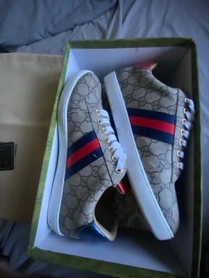 Gucci sneakers med GG-mönster - Snygga Gucci sneakers i beige canvas med GG-monogram, blå och röda ränder på sidan samt metallicblå och röd häl i ormskinnsmönster. Vita snören och gummisula. Perfekt för dig som vill sticka ut med klassiska men ändå unika detalj.
