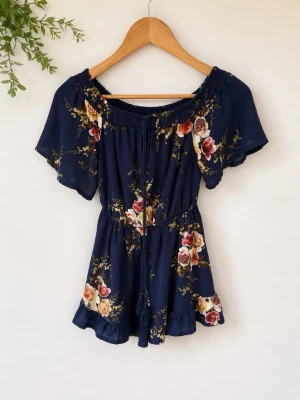 Blommig marinblå jumpsuit Parisian - Supersöt marinblå jumpsuit från Parisian med blommigt mönster i rosa, gult och grönt. Off shoulder-modell med resår upptill, korta vida ärmar och volangdetalj nertill. Perfekt för en trendig och somrig look.