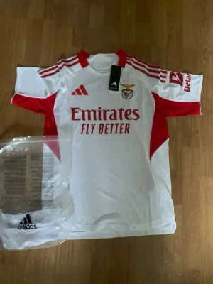 Fotbollströja  - Säljer en officiell Benfica matchtröja från Adidas i vitt med röda detaljer och klubbmärke på bröstet. Tröjan har röda ränder på axlarna, sponsortryck framtill och klassisk sportig passform. Perfekt för dig som älskar fotboll och vill stötta ditt lag.