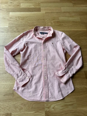 Ljusrosa skjorta från Ralph Lauren - Ljusrosa skjorta från Ralph Lauren i modellen Custom Fit. Skjortan är i mjuk bomull, har klassisk krage, knappar framtill och lång ärm. Broderad logga på bröstet ger en clean och stilren vibe.