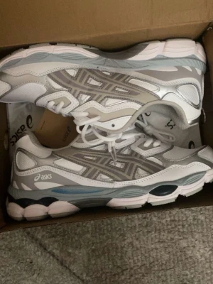 Vita och silver Asics sneakers - Snygga vita och silverfärgade Asics sneakers med mesh och syntetdetaljer. Skorna har en chunky sula med blå och svarta inslag samt klassiska Asics-loggor på sidan. Perfekta för dig som gillar sportig och trendig stil.