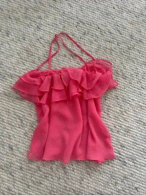 Rosa volang topp med tunna axelband - Säljer en söt rosa topp med tunna axelband och volangdetaljer upptill. Toppen är ärmlös och har en luftig, flowy passform i ett lätt och transparent material som känns perfekt för varma dagar. Supersnygg att styla till jeans eller kjol!