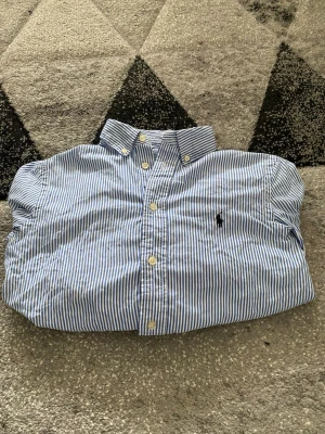 Blårandig skjorta från Polo Ralph Lauren - Säljer en klassisk blårandig skjorta från Polo Ralph Lauren i slim fit. Skjortan har vit knappstängning, button-down krage och den ikoniska broderade loggan på bröstet. Tillverkad i mjuk bomull och passar perfekt till en clean och stilren look.