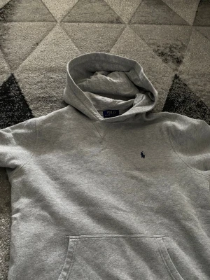 Grå hoodie från Polo Ralph Lauren - Säljer en klassisk grå hoodie från Polo Ralph Lauren med huva och magficka. Tröjan har det ikoniska lilla mörkblå logobroderiet på bröstet och är tillverkad i mjukt sweatshirtmaterial. Perfekt för en avslappnad och stilren look.
