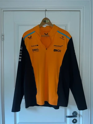 McLaren F1 långärmad tröja orange/svart - Snygg långärmad tröja från McLaren F1 Team i orange och svart med blå detaljer. Halv dragkedja framtill, tryckta loggor från sponsorer och teamet både fram och bak. Perfekt för motorsportfans som vill sticka ut med stil. Den är från officiella merch från mclaren, köpt i Belgien på SPA.  Använt en gång, helt ny men utan pris lapp.