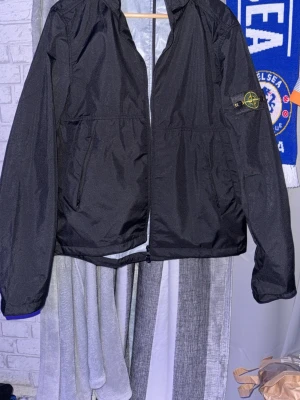 Svart vindjacka från Stone Island - Snygg svart vindjacka från Stone Island med huva och dragkedja framtill. Jackan har två snedställda fickor med dragkedja och den klassiska Stone Island-loggan på vänster ärm. Tillverkad i lätt och tåligt material, perfekt för blåsiga dagar. Storlek M men passar även S skulle jag säga