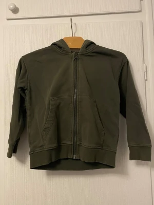 Mörkgrön hoodie med dragkedja - En enkel och stilren mörkgrön hoodie med dragkedja framtill och känguruficka. Tröjan har huva och ribbade muddar vid ärmslut och nederkant. Perfekt för en avslappnad look och passar till det mesta. Tillverkad i mjukt bomullsmaterial.