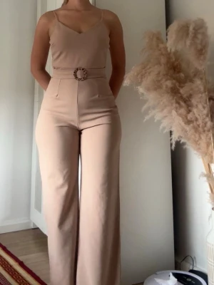 Beige jumpsuit med bälte - Snygg beige jumpsuit med smala axelband och v-ringning. Vida ben och markerad midja med bälte som har ett leopardmönstrat spänne. Tillverkad i ett mjukt, stretchigt tyg som sitter bekvämt och ger en clean look.