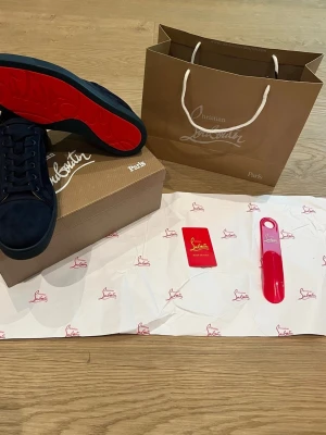 Marinblå sneakers från Christian Louboutin - Snygga marinblå sneakers från Christian Louboutin med ikoniska röda sulor och diskret logga på sidan. Skorna har klassisk låg profil, rund tå och är tillverkade i mocka med matchande snörning. Perfekta för dig som vill sticka ut med exklusiv stil.