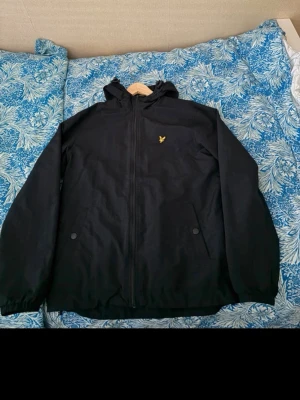 Svart vindjacka med huva - Svart vindjacka med dragkedja och huva. Jackan har två framfickor med knappar och ett litet gult broderat emblem på bröstet. Resår vid ärmslut och nederkant. Perfekt för blåsiga dagar men generellt passar den till både vinter, höst och vår när det är lite kylligt