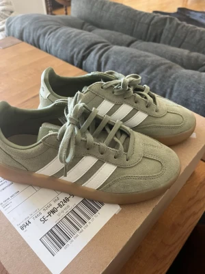 Adidas sneakers i grön mocka - Snygga Adidas sneakers i ljusgrön mocka med klassiska vita ränder på sidorna och gummisula. Skorna har lågt skaft och rund tå, perfekta för en avslappnad streetstyle-look. Skosnören i matchande grön färg och diskret Adidas-logga på hälen.
