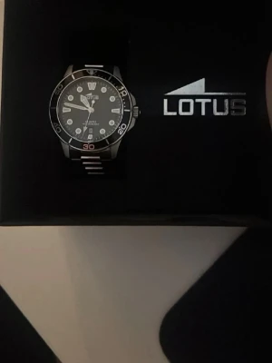 Svart och silver Lotus herrklocka - Stilren herrklocka från Lotus med svart urtavla och silverfärgade detaljer. Klockan har en rund boett, tydliga siffror och markeringar samt en länk i silverfärgad metall. Extra länkbitar medföljer för justering av armbandet.