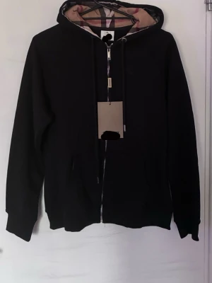 Svart hoodie med dragkedja och huva - Snygg svart hoodie med dragkedja och klassisk huva. Insidan av huvan har ett beige-rutigt mönster som ger en unik touch. Hoodien har fickor framtill och ribbade muddar vid ärmar och nederkant. Perfekt för en chill och avslappnad stil. Storleken är enligt etiketten xxl men den är mycket mindre så skulle nog säga xs,s,m eller L, den är helt ny endast testad, endast seriösa köpare om ni vill diskutera pris… alltså om ni är intresserade så kan ni skicka prisförslag 
