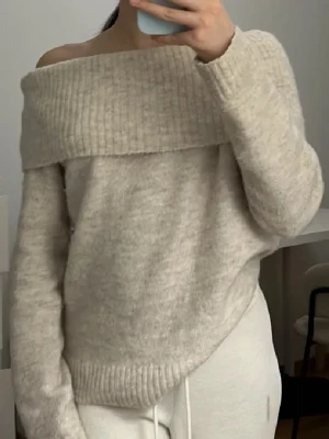 Beige off shoulder stickad tröja - Säljer en beige stickad tröja med bred off shoulder-krage och långa ärmar. Tröjan har en mjuk, fluffig känsla och ribbstickade detaljer vid krage och nederkant. Perfekt för en avslappnad och trendig look.