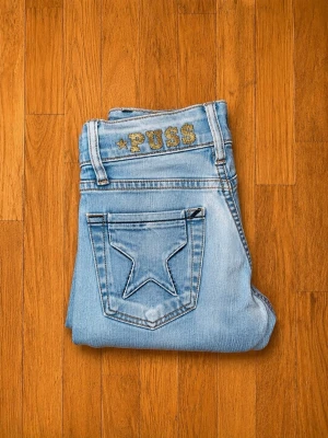Puss & Kram Y2K jeans – ljusblå låg midja med stjärnfickor - Märke: Puss & Kram Storlek: W24 / motsvarar cirka XS (EU 34) Material: 98% bomull, 2% elastan Färg: Ljusblå denim Skick: Mycket bra begagnat skick  Mått (ca):  * Midja: 31 cm platt mätt (+ stretch) * Innerbenslängd: 80 cm