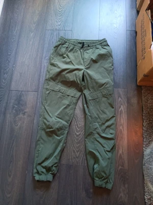 Olivgröna cargobyxor med resår - Säljer ett par olivgröna cargobyxor med resår i midjan och vid bensluten. Byxorna har flera fickor och är tillverkade i ett lätt, slitstarkt material som passar perfekt för en avslappnad streetwear-look.