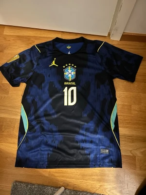 Brasilien Neymar JR matchtröja blå 2026 - Snygg blå och svart Brasilien matchtröja med Neymar JR och nummer 10 på ryggen. Tröjan har gula och gröna detaljer, rund hals och korta ärmar. Tillverkad i ett lätt och ventilerande material, perfekt för fotboll eller träning.