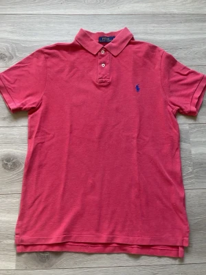 Rosa pikétröja från Polo Ralph Lauren - Klassisk rosa pikétröja från Polo Ralph Lauren med blå broderad logga på bröstet. Tröjan har krage, två knappar framtill och korta ärmar. Perfekt till jeans eller shorts för en avslappnad men snygg look. Röd rosa piké tröja i super skick passar storlek M 