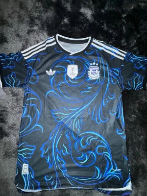 Argentina Adidas fotbollströja svart/blå - Snygg svart fotbollströja från Adidas med blått och lila mönster, vita detaljer och tre ränder på axlarna. Bröstet har FIFA World Champions 2022-märke och Argentinas landslagsemblem. Perfekt för dig som älskar fotboll och vill sticka ut på planen.