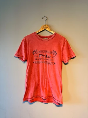 Röd t-shirt från Polo Ralph Lauren - Säljer en röd t-shirt från Polo Ralph Lauren med svart tryck framtill. T-shirten har rund halsringning och korta ärmar. Materialet känns mjukt och skönt, perfekt för en avslappnad stil.