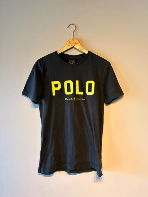 Svart POLO t-shirt från Ralph Lauren - Snygg svart t-shirt från Ralph Lauren med stor gul POLO-logga och text på bröstet. Klassisk rund hals och korta ärmar. Perfekt till jeans eller shorts för en clean och sportig look.