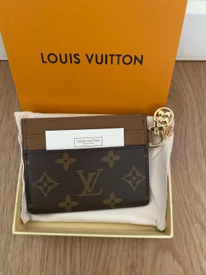Louis Vuitton monogram korthållare - Snygg korthållare från Louis Vuitton i klassiskt brunt monogrammönster med guldiga detaljer och charm. Tillverkad i läder med plats för flera kort. Perfekt accessoar för dig som gillar lyxiga detaljer och stilren design.