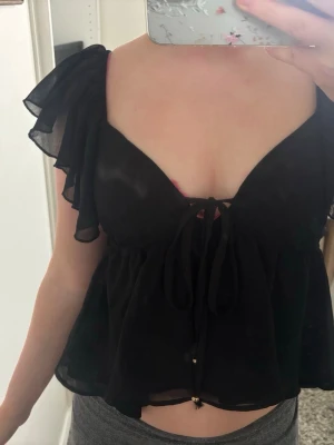 Svart blus med volangärmar och knyt - Supersöt svart blus med volangärmar och djup v-ringning. Toppen har knyt framtill och är lätt och luftig i ett tunt material, perfekt för varma dagar. Passformen är loose och toppen är något croppad. Perfekt att styla med jeans eller kjol. Använd 1 gång men är tyvärr för liten