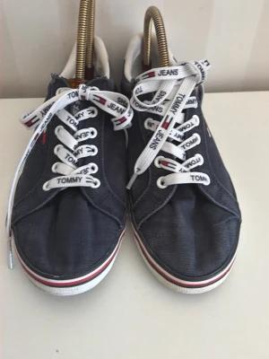 Marinblå Tommy Jeans sneakers - Snygga marinblå sneakers från Tommy Jeans med vita snören där märkesnamnet står tryckt. Skorna har lågt skaft, vit sula med röd och blå rand samt klassisk design i textilmaterial. Perfekta för en avslappnad och trendig look.