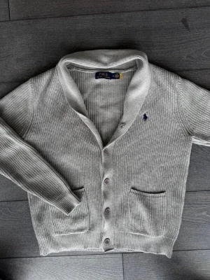 Grå kofta från Polo Ralph Lauren - Snygg grå kofta från Polo Ralph Lauren med ribbstickat material, knäppning framtill och två fickor. Klassisk design med broderad logga på bröstet. Perfekt för lager-på-lager och enkel att matcha med olika outfits.