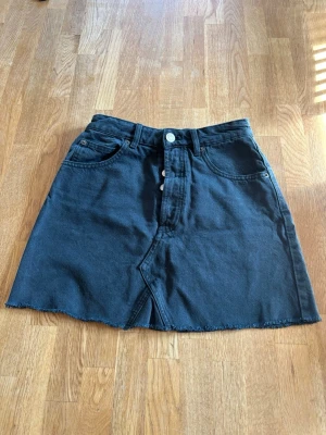 Svart jeanskjol från Zara - Säljer en svart jeanskjol från Zara i klassisk A-linjeform. Kjolen har rå kant nertill, knappstängning framtill samt fickor både fram och bak. Perfekt för dig som gillar en enkel men snygg stil.