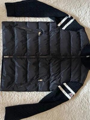 Svart pufferjacka från Moncler - Snygg svart pufferjacka från Moncler med stickade ärmar och två vita ränder på axlarna. Jackan har två snedställda dragkedjefickor framtill och Moncler-logga på ena ärmen. Kroppen är vadderad medan ärmar och rygg är kabelstickade.