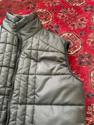 Svart dunväst från Louis Vuitton - Stilren svart dunväst från Louis Vuitton med diskret LV-logga på bröstet. Västen har hög krage, två dragkedjefickor och quiltat mönster. Perfekt för lager-på-lager och ger en lyxig touch till din outfit.